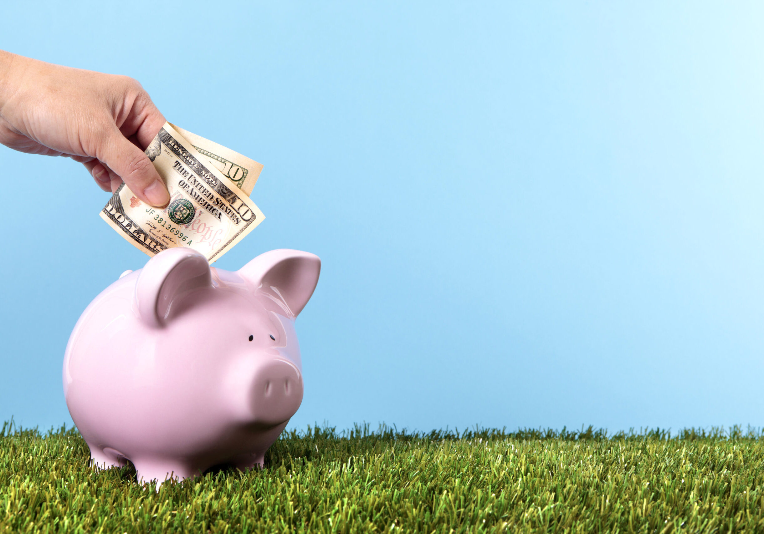 Hand putting a ten dollar bill into a pink piggy bank, with grass and blue sky